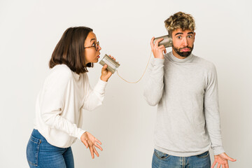 Young hispanic friends talking through a tin can system shrugs shoulders and open eyes confused.