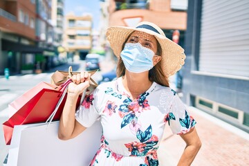 Middle age hispanic woman wearing medical mask holding shopping bags at the city