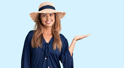 Middle age hispanic woman wearing summer hat smiling cheerful presenting and pointing with palm of hand looking at the camera.