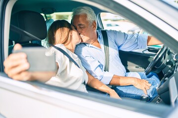 Middle age couple in love sitting inside the car going for a trip taking a selfie picture with smartphone kissing