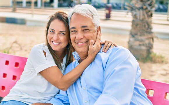 Middle age hispanic couple smiling happy and hugging sitting on bench at the beach.