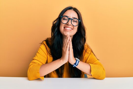 Beautiful brunette young woman wearing glasses and casual clothes sitting on the table praying with hands together asking for forgiveness smiling confident.