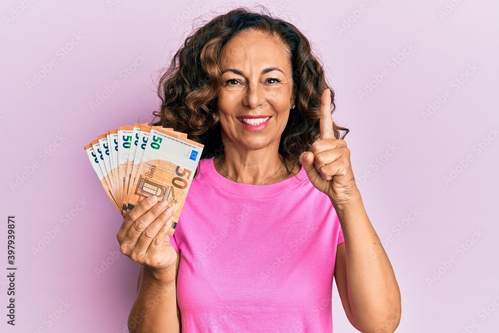 Wall mural Middle age hispanic woman holding euro banknotes smiling with an idea or question pointing finger with happy face, number one