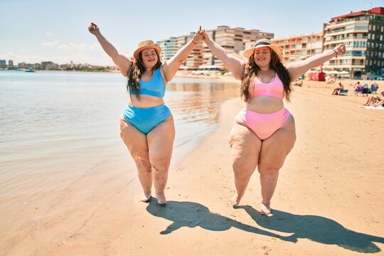 Two plus size overweight sisters twins women happy and proud of their bodies walking at the beach on summer holidays