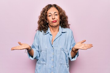 Middle age beautiful woman wearing casual denim shirt standing over pink background clueless and confused with open arms, no idea concept.