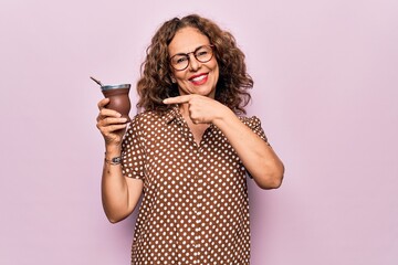 Middle age beautiful woman drinking cup of mate over isolated pink background smiling happy...