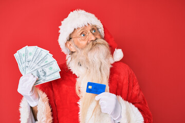 Old senior man wearing santa claus costume holding dollars and credit card smiling looking to the side and staring away thinking.