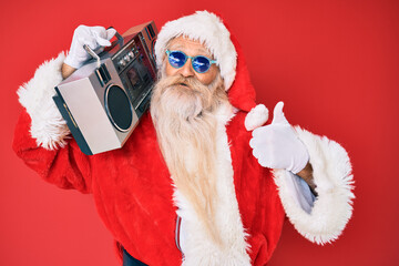 Old senior man wearing santa claus costume and boombox smiling happy and positive, thumb up doing excellent and approval sign