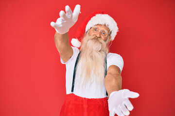 Old senior man with grey hair and long beard wearing white t-shirt and santa claus costume looking at the camera smiling with open arms for hug. cheerful expression embracing happiness.