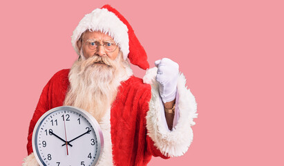 Old senior man with grey hair and long beard wearing santa claus costume holding clock annoyed and frustrated shouting with anger, yelling crazy with anger and hand raised