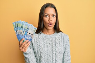 Beautiful hispanic woman holding south african 100 rand banknotes scared and amazed with open mouth for surprise, disbelief face