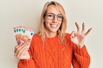 Beautiful blonde woman holding 10 euro banknotes doing ok sign with fingers, smiling friendly...