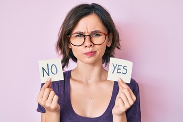 Young beautiful girl holding yes and no reminder skeptic and nervous, frowning upset because of...