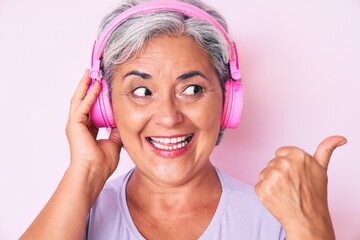 Senior hispanic woman listening to music using headphones pointing thumb up to the side smiling happy with open mouth