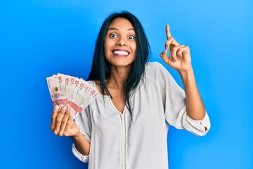 Obraz premium Young african american woman holding 10 colombian pesos banknotes smiling amazed and surprised and pointing up with fingers and raised arms.
