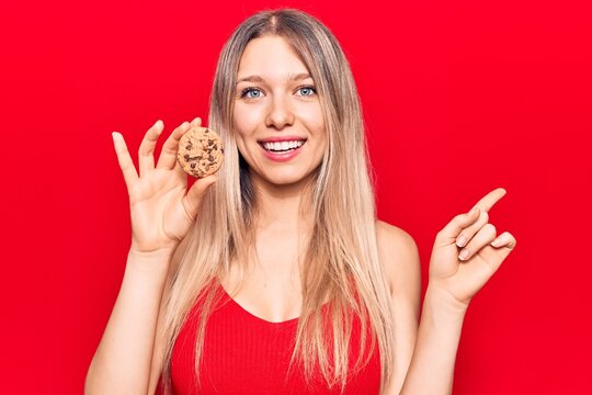 Young beautiful blonde woman holding cookie smiling happy pointing with hand and finger to the side