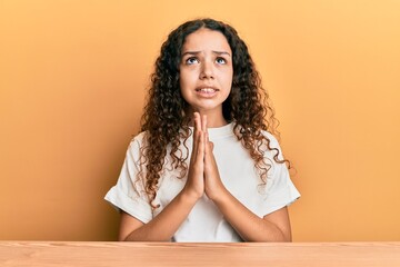 Teenager hispanic girl wearing casual clothes sitting on the table begging and praying with hands...