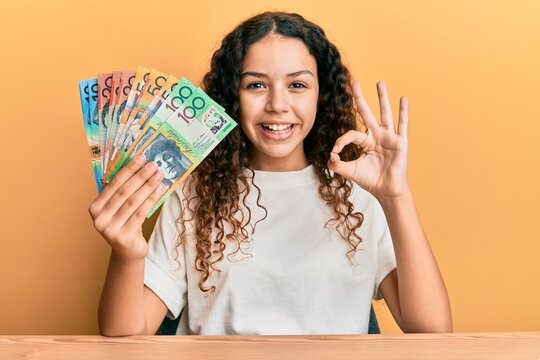 Teenager hispanic girl holding australian dollars doing ok sign with fingers, smiling friendly gesturing excellent symbol