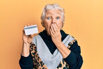 Senior grey-haired woman holding credit card covering mouth with hand, shocked and afraid for...