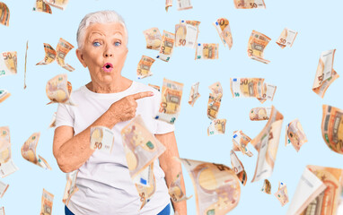 Senior beautiful woman with blue eyes and grey hair wearing casual white tshirt surprised pointing with finger to the side, open mouth amazed expression.
