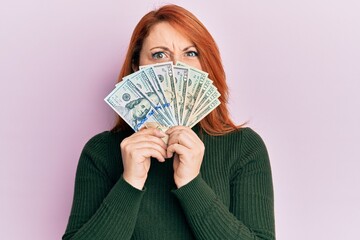 Beautiful redhead woman holding dollars close to face clueless and confused expression. doubt concept.