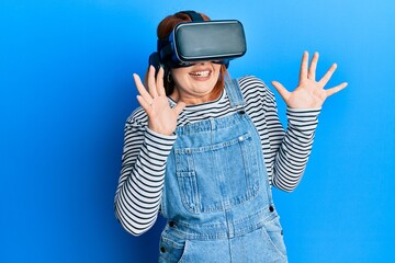 Young redhead woman scared using 3d virtual glasses standing over isolated blue background.