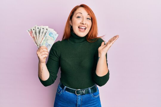 Beautiful redhead woman holding united kingdom pounds celebrating achievement with happy smile and winner expression with raised hand