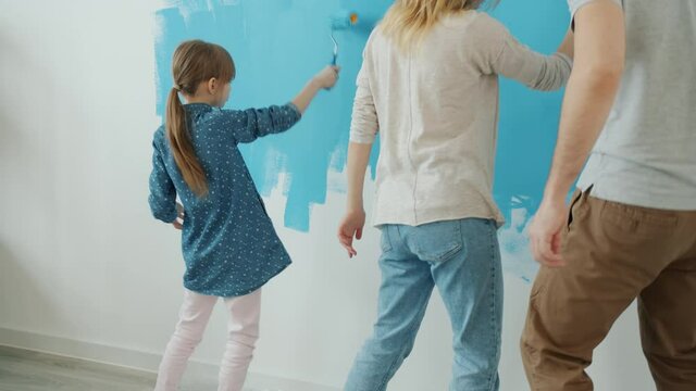 Back view of happy young couple with kid painting wall at home and dancing renovating apartment together. Family life and accommodation concept.