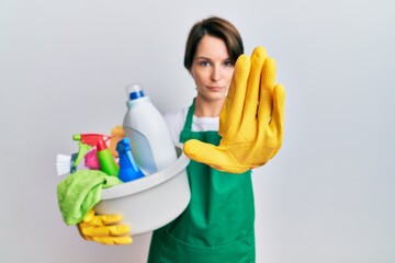 Young brunette woman with short hair wearing apron holding cleaning products doing stop sing with...