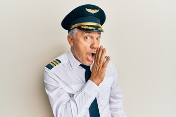 Handsome middle age mature man wearing airplane pilot uniform hand on mouth telling secret rumor, whispering malicious talk conversation