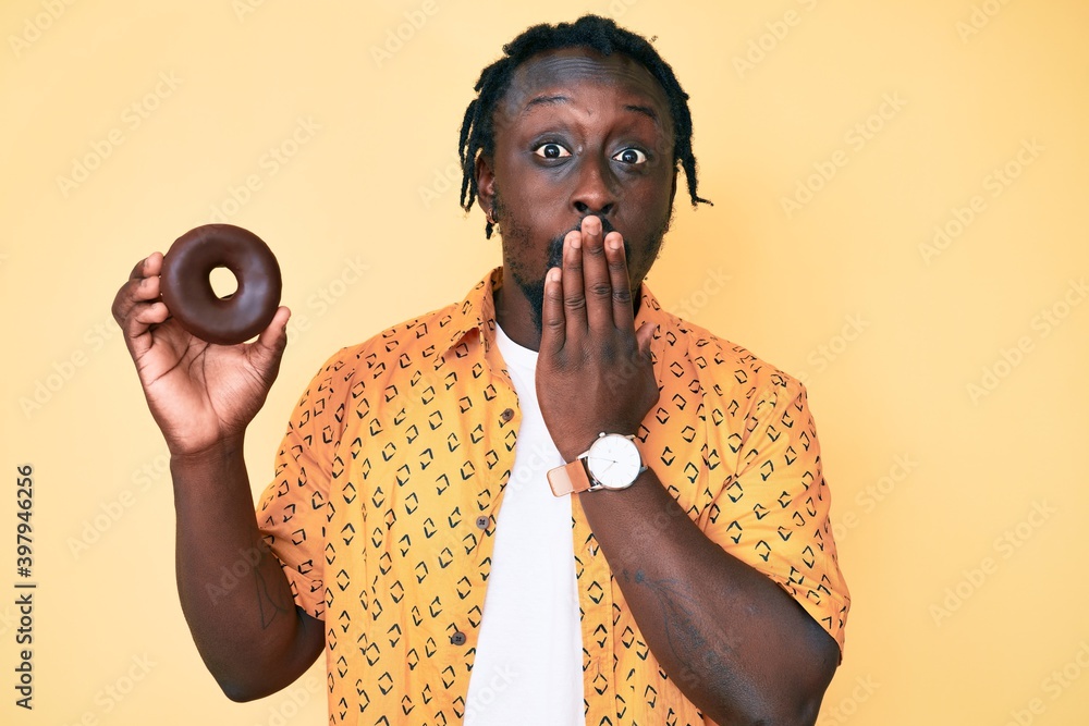 Canvas Prints Young african american man with braids holding donut covering mouth with hand, shocked and afraid for mistake. surprised expression