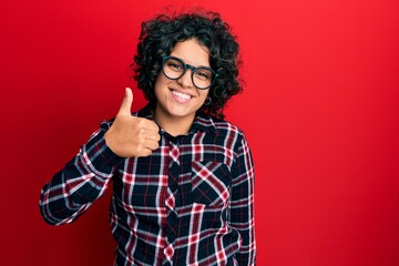 Young hispanic woman with curly hair wearing casual clothes and glasses doing happy thumbs up gesture with hand. approving expression looking at the camera showing success.