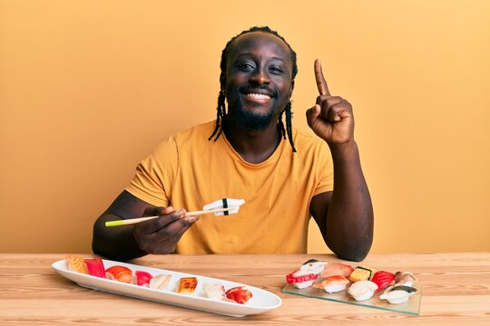 Handsome young black man eating sushi sitting on the table smiling with an idea or question pointing finger with happy face, number one