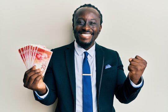 Handsome young black man wearing business suit holding 20 shekels banknotes screaming proud, celebrating victory and success very excited with raised arm