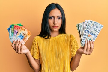 Young brunette woman holding bunch of dollars and swiss francs banknotes relaxed with serious...
