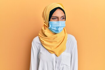 Young brunette arab woman wearing traditional islamic hijab and medical mask looking away to side with smile on face, natural expression. laughing confident.