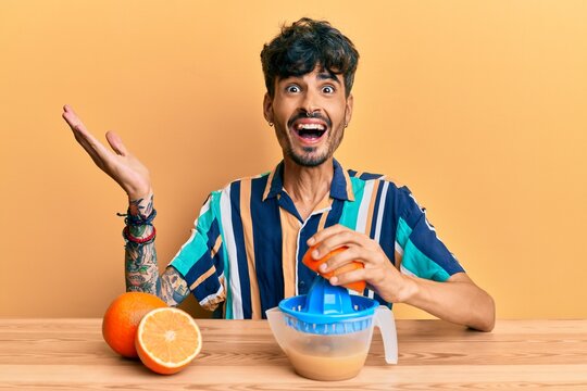 Young Hispanic Man Sitting On The Table Using Juicer Celebrating Victory With Happy Smile And Winner Expression With Raised Hands