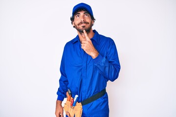 Handsome young man with curly hair and bear weaing handyman uniform thinking concentrated about...