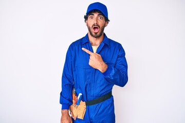 Handsome young man with curly hair and bear weaing handyman uniform surprised pointing with finger to the side, open mouth amazed expression.