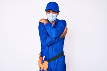 Handsome young man with curly hair and bear wearing handyman uniform and covid-19 safety mask hugging oneself happy and positive, smiling confident. self love and self care