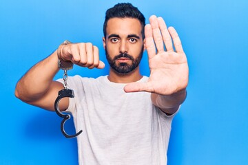 Young hispanic man holding prisoner handcuffs with open hand doing stop sign with serious and confident expression, defense gesture