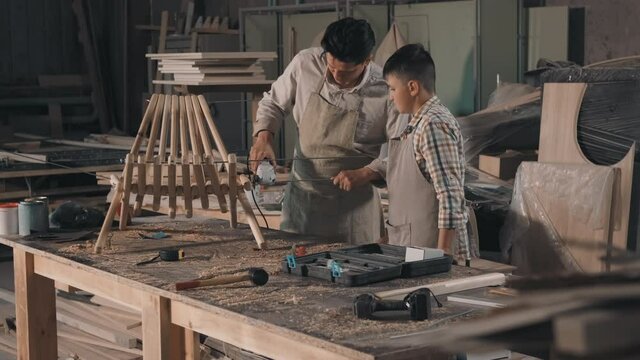 Medium shot of happy father and son spending time together in carpentry workshop constructing new wooden deck chair sawing wooden planks