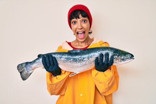 Beautiful brunettte fisher woman wearing raincoat holding fresh salmon celebrating crazy and amazed for success with open eyes screaming excited.