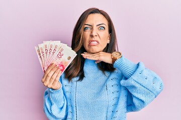 Young brunette woman holding 20 israel shekels banknotes cutting throat with hand as knife, threaten aggression with furious violence