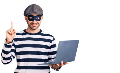 Young handsome man wearing burglar mask using laptop surprised with an idea or question pointing finger with happy face, number one