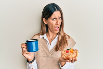 Brunette young woman drinking coffee and eating pastry in shock face, looking skeptical and sarcastic, surprised with open mouth