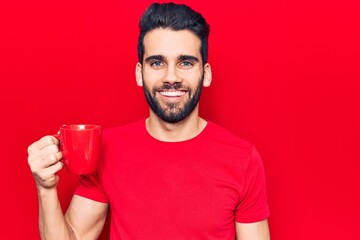 Young handsome man with beard drinking mug of coffee looking positive and happy standing and smiling with a confident smile showing teeth