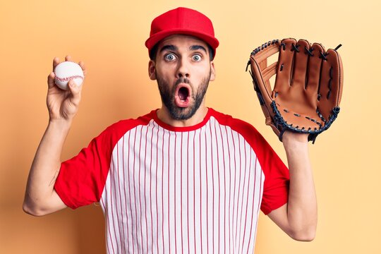 Young handsome man with beard playing baseball using ball and glove scared and amazed with open mouth for surprise, disbelief face