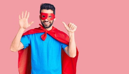 Young handsome man with beard wearing super hero costume showing and pointing up with fingers number six while smiling confident and happy.