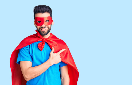 Young handsome man with beard wearing super hero costume cheerful with a smile of face pointing with hand and finger up to the side with happy and natural expression on face